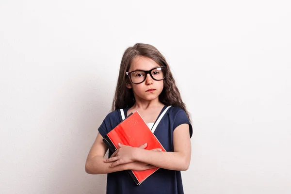 Маленька школярка в окулярах і уніформі стоїть в студії, тримаючи блокнот . — стокове фото