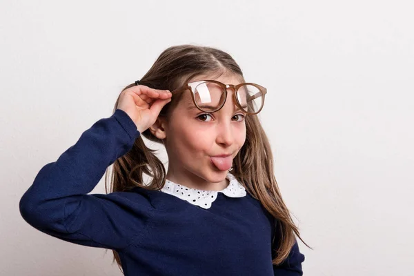 Malá drzá školačka s brýlemi a uniforma v ateliéru, trčí jako jazyk. — Stock fotografie