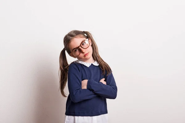 Una pequeña colegiala triste con gafas y uniforme de pie en un estudio, brazos cruzados . —  Fotos de Stock