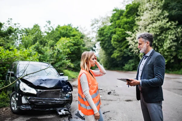 An insurance agent talking to a woman driver by the car on the road after an accident. — Stock Photo, Image