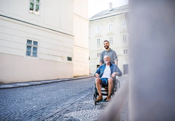 Dorosłego syna z Starszy ojciec na wózku inwalidzkim na spacer w mieście. — Zdjęcie stockowe