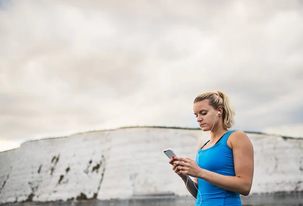 Sportig kvinna löpare med hörlurar och smartphone stående på stranden. — Stockfoto
