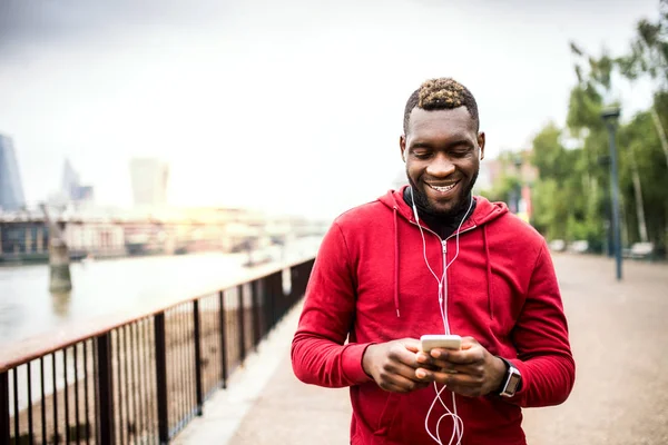 Fiatal sportos fekete férfi futó a hídon kívül a városban, használ smartphone. — Stock Fotó