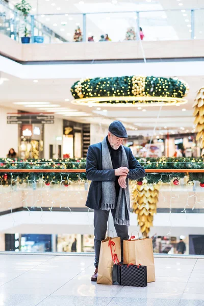 Senior man met zakken staan in een winkelcentrum kerst tijd, de tijd controleren. — Stockfoto