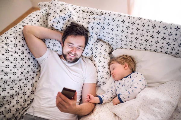 Pai com smartphone e um menino adormecido na cama em casa . — Fotografia de Stock