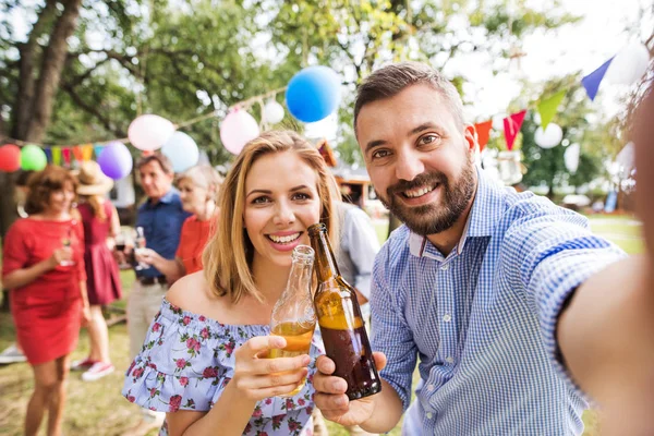 Ungt par tar selfie på en fest utanför i trädgården, spottar flaskor. — Stockfoto