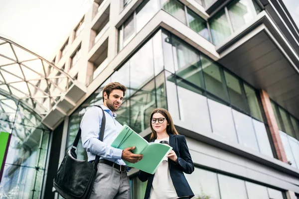 A young businessman and businesswoman standing in front of a building, looking at notes. — Stock Photo, Image