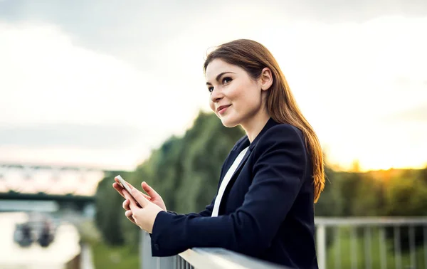 A young businesswoman standing on the river bank, using smartphone. — Stock Photo, Image