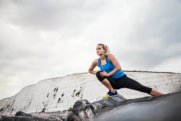 ストレッチ、外岩の上に立ってイヤホンでスポーティな女性ランナー. — ストック写真