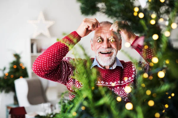 En äldre man står vid julgran, sätta bollar framför ögonen. — Stockfoto