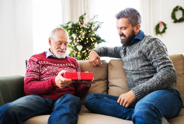 Um pai sênior e um filho adulto com um presente sentado em um sofá na época do Natal . — Fotografia de Stock