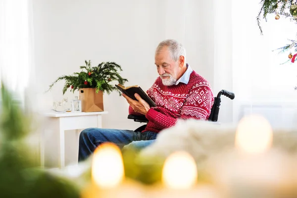 Un aîné en fauteuil roulant lisant un livre à la maison à Noël . — Photo