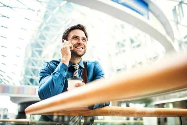 Ein junger Geschäftsmann mit Smartphone steht in einem modernen Gebäude und telefoniert. — Stockfoto
