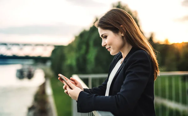 Een jonge zakenvrouw permanent aan de rivieroever, met behulp van smartphone. — Stockfoto