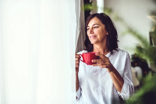 A senior woman with a cup standing by the window at home at Christmas time. — Stock Photo, Image