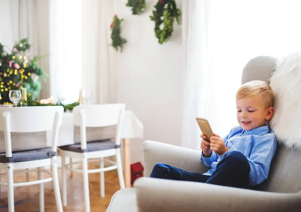 En liten pojke med smartphone som sitter på en fåtölj hemma i juletid. — Stockfoto
