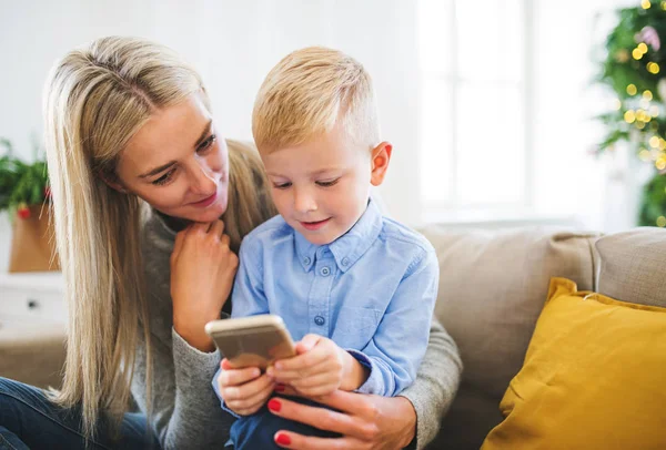 Una mamma e un bambino con smartphone seduti su un divano a casa nel periodo natalizio . — Foto Stock