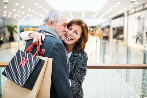 Heureux couple aîné avec des sacs en papier dans le centre commercial, câlin . — Photo