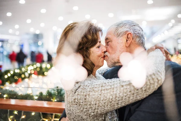 Een gelukkige senior paar permanent in shopping center, het zoenen. — Stockfoto