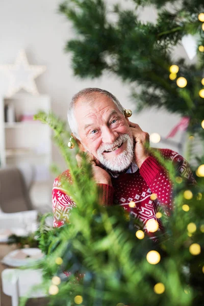 A senior man standing by Christmas tree, putting ball ornaments on his ears. — Stock Photo, Image