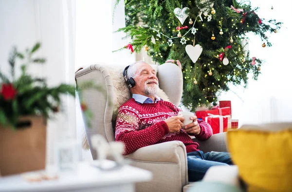 Ett porträtt av äldre man med hörlurar som sitter på fåtölj hemma i juletid. — Stockfoto