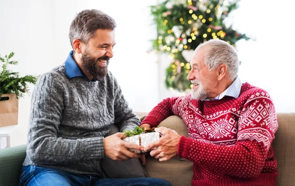 Um pai sênior e um filho adulto com um presente sentado em um sofá na época do Natal . — Fotografia de Stock