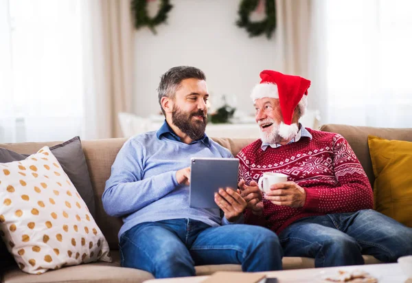 A senior father and adult son with tablet sitting on a sofa at home at Christmas time. — Stock Photo, Image