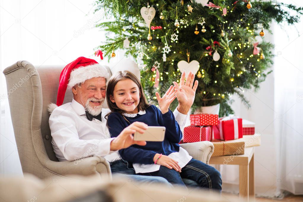 A small girl and her grandfather with Santa hat taking selfie with smartphone at Christmas time.