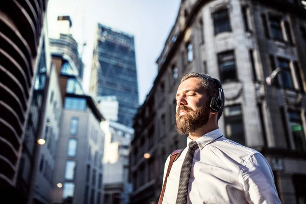 Hipster businessman with headphones standing on the street in city. Copy space.