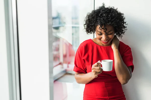 Un primer plano de una mujer de pie junto a la ventana sosteniendo la taza de café . — Foto de Stock