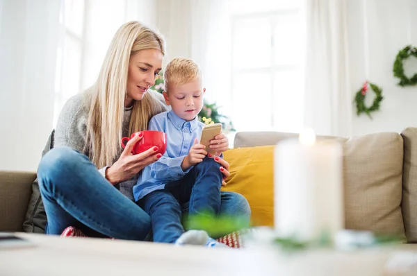 Una mamma e un bambino con smartphone seduti su un divano a casa nel periodo natalizio . — Foto Stock