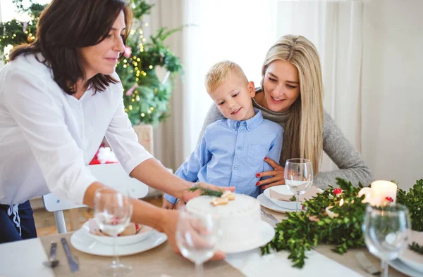 Um menino pequeno com a mãe olhando para a avó colocando um bolo na mesa na época do Natal . — Fotografia de Stock