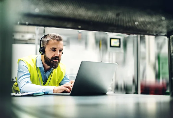 Um engenheiro industrial com fone de ouvido e laptop em uma fábrica, trabalhando . — Fotografia de Stock