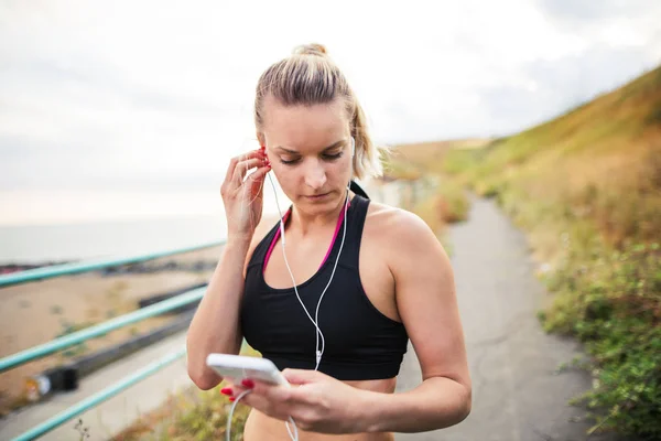 Sportieve jongedame loper met oortelefoons en smartphone staande aan de kust. — Stockfoto