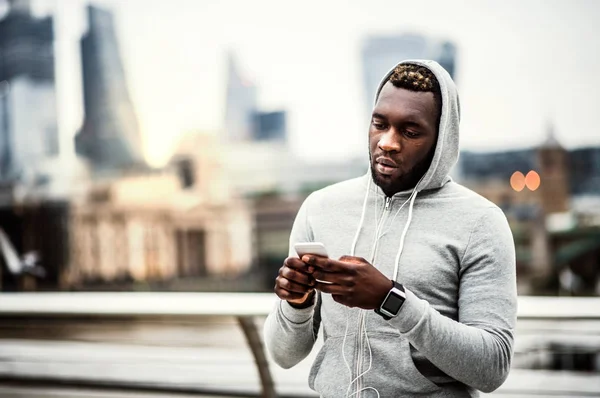 Black man runner with smart watch and smartphone on the bridge in a city, resting.