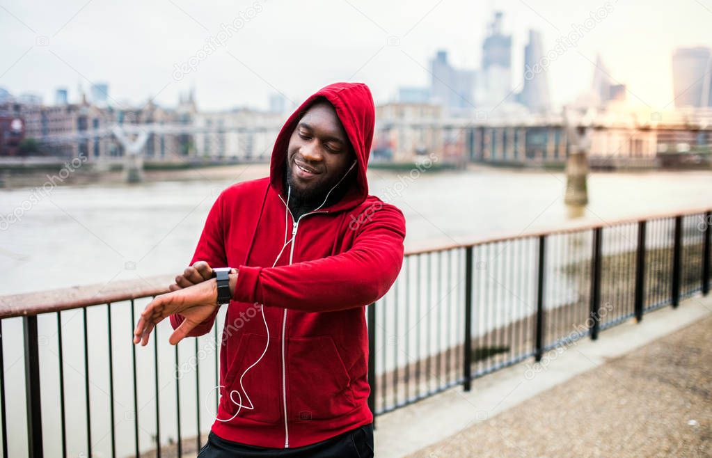 Young sporty black man runner on the bridge outside in a city, setting smartwatch.