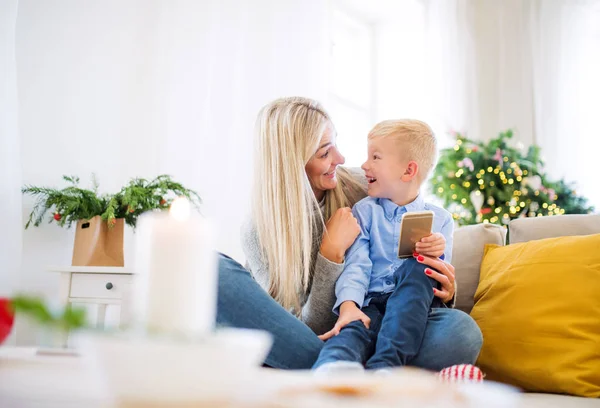 Uma mãe e um menino pequeno com smartphone sentado em um sofá em casa na época do Natal . — Fotografia de Stock