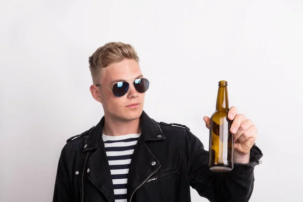 Een zelfverzekerde jonge man met zonnebril in een studio, bedrijf leeg bierflesje. — Stockfoto