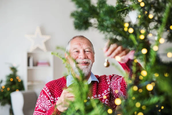 Um homem sênior em pé junto à árvore de Natal, segurando bolas ornamentos . — Fotografia de Stock