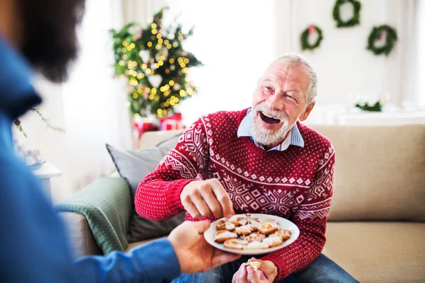 Um homem dando biscoitos para seu pai sênior em casa na época do Natal . — Fotografia de Stock