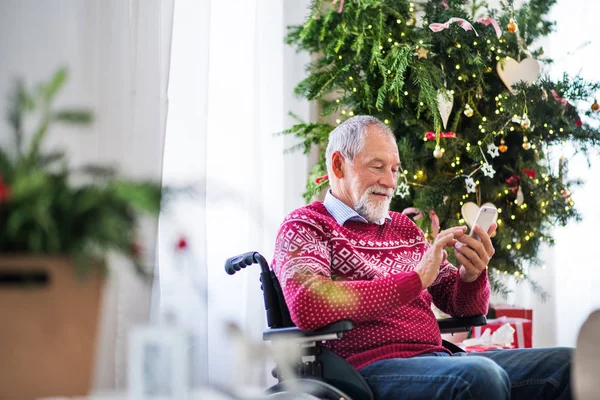 Un homme âgé en fauteuil roulant avec téléphone portable à la maison à la période de Noël, textos . — Photo