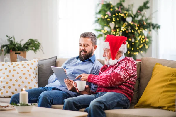 Um pai sênior e filho adulto com tablet sentado em um sofá em casa na época do Natal . — Fotografia de Stock