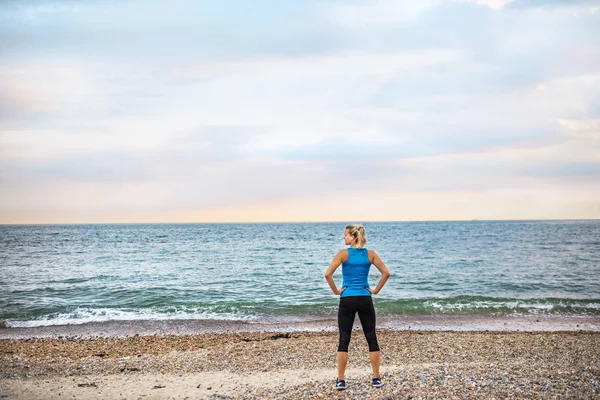 Vista posteriore di giovane donna sportiva corridore in abbigliamento sportivo blu in piedi sulla spiaggia . — Foto Stock