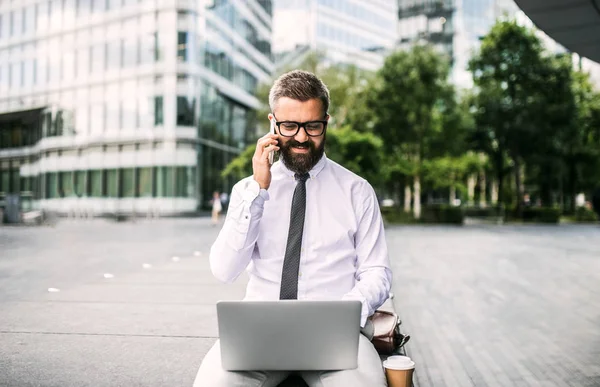 Uomo d'affari hipster con laptop e smartphone seduto all'aperto in città . — Foto Stock