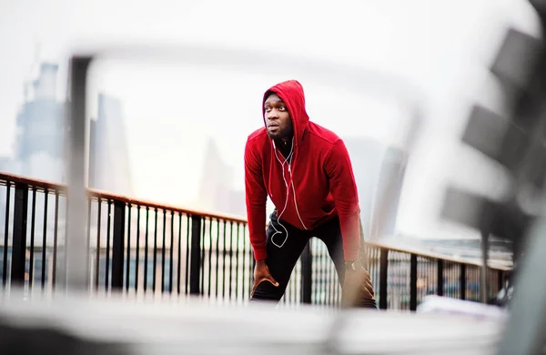 A black man runner with earphones and hood on his head in a city, resting.