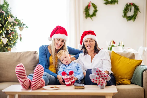 Een kleine jongen met moeder en grootmoeder met KERSTMUTS thuis bij Kerstmis. — Stockfoto