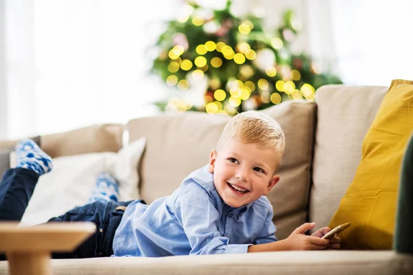 En liten pojke med smartphone liggande på en soffa hemma i juletid. — Stockfoto