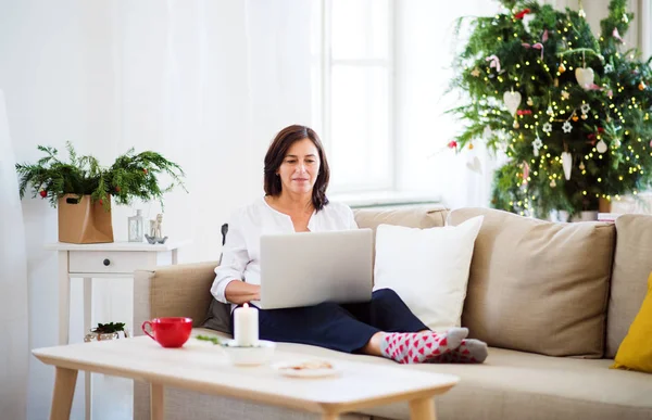 Eine Seniorin mit Laptop sitzt zur Weihnachtszeit zu Hause auf dem Sofa. — Stockfoto