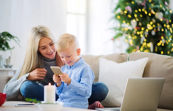 Una madre y un niño pequeño con teléfono inteligente sentado en un sofá en casa en Navidad . — Foto de Stock