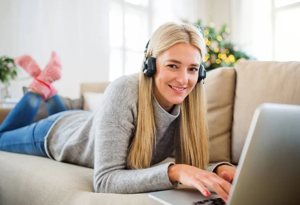 Uma jovem mulher com fones de ouvido e laptop em casa na época do Natal . — Fotografia de Stock